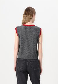 Pull sans manches en tricot gris avec motif en relief et bords côtelés. Accent de col rouge. Associé à un jean noir taille haute.