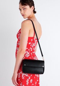 Borsa a tracolla nera in pelle con forma rettangolare, due dettagli con bottone a pressione, una lunga tracolla regolabile e una texture liscia.