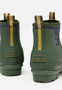 Zielone gumowe botki za kostkę o teksturowanej powierzchni, z złotymi akcentami i paskowymi tkaninowymi uchwytami. Posiadają niebieską kratkę wewnątrz oraz detal z logo.