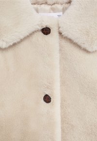 Beige kunstpelsfrakke med en blød tekstur, der har en spidse krave og brune knapdetaljer. Glat foer synligt ved kraven.