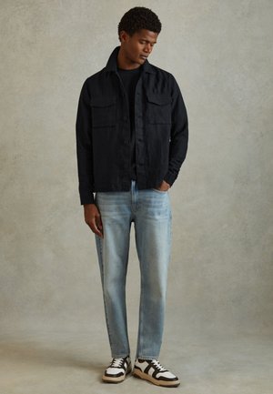 Mand iført sort jakke med knapper, sort skjorte, lyseblå jeans og hvide og sorte sneakers, stående mod beige baggrund.