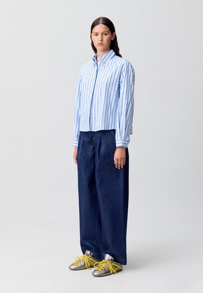 Modelo de pie contra un fondo liso, vistiendo una camisa de rayas azul y blanca de gran tamaño, pantalones de mezclilla anchos y zapatillas plateadas con cordones amarillos.