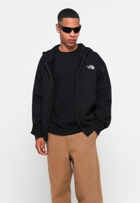 Black zip-up hoodie with logo, layered over a black t-shirt. Brown relaxed-fit pants. Sunglasses complete the outfit. Smooth fabric texture.