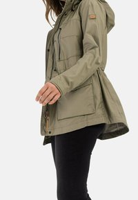 Femme portant une veste à capuche vert olive clair avec fermeture éclair et grandes poches avant, associée à un pantalon noir, vue de profil.