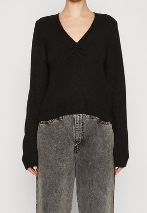 Personne portant un pull noir à col en V manches longues et un jean gris délavé taille haute sur un fond uni.