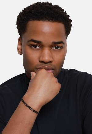 Bracciale in catena nera in metallo con segmenti collegati, indossato al polso, su una camicia nera, che mette in risalto la texture e i dettagli del design.