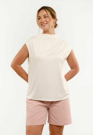 Kobieta o blond włosach związanych w niski kok, ubrana w białą bluzkę bez rękawów i jasnoróżowe krótkie spodenki, uśmiecha się i patrzy na bok.