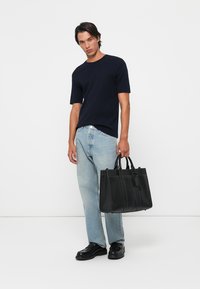 Svart lær tote-veske med to topphåndtak, strukturert design og kontrastfargede sømdetaljer, holdt av en person i en marineblå skjorte og lyse jeans.