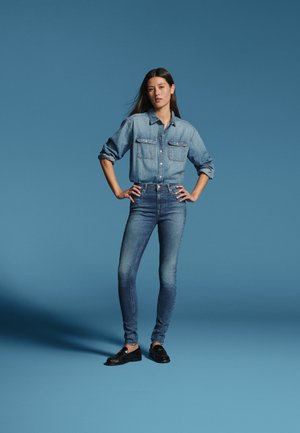 Kvinde stående med hænderne på hoften iført en denimskjorte og jeans mod en ensfarvet blå baggrund.
