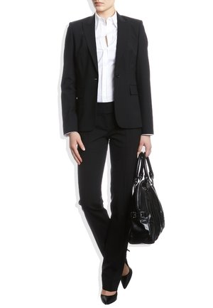 Zwart op maat gemaakt pak met een getailleerde blazer over een witte blouse, gecombineerd met zwarte broek en puntige hakken. Een zwarte handtas wordt vastgehouden.