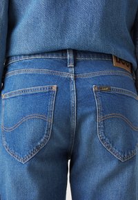 Jeans od trapera u srednje plavoj boji s narančastim šavovima, imaju dvije stražnje džepove s zaobljenim rubovima i označenu kožnu oznaku na pojasu.