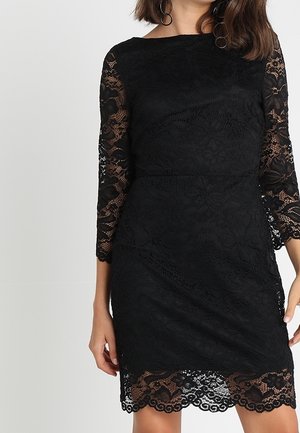 Frau trägt ein figurbetontes schwarzes Spitzenkleid mit dreiviertel Ärmeln und gewelltem Saum vor einem schlichten Hintergrund.