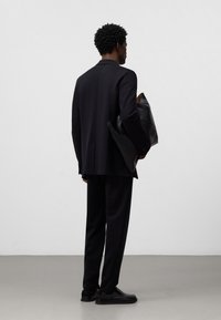 Abito nero su misura con una sola apertura, abbinato a una borsa nera texturizzata. Scarpe lisce e sfondo neutro completano il look.