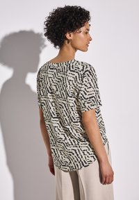Top beige a maniche corte con motivo astratto nero, orlo arrotondato e vestibilità comoda, abbinato a pantaloni larghi di colore chiaro.