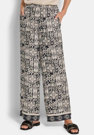 Weit geschnittene Hose mit schwarzen und beigefarbenen, komplizierten geometrischen und floralen Mustern, getragen mit schwarzen Slide-Sandalen und Händen in den Taschen.