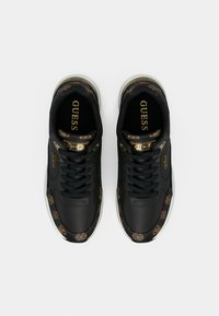 Paire de baskets noires Guess avec des accents marron à motifs, des détails de logo dorés et des semelles blanches, vues de dessus sur un fond blanc.
