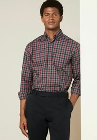 Heren lange mouw geruit shirt in rood, marineblauw en grijze tinten, met een button-down kraag en opgerolde manchetten, gecombineerd met donkere broeken.