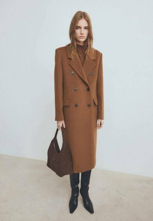 Brown wool-blend double-breasted coat with large lapels, black buttons, and two side pockets, paired with sleek black leather boots and a dark bag.