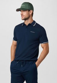 Man met een groene pet en een marineblauw Hackett polohemd, bijpassende marineblauwe broek met trekkoord, handen in de zakken, kijkt naar links.