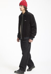 Zwarte corduroy jas met een kraag, ritssluiting aan de voorkant en zijkanten. Draag het over een grijze T-shirt en combineer het met zwarte broek en schoenen. Rode beanie.