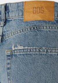 Jeans de ganga azul claro com detalhe de bolso desgastado e etiqueta de couro castanho em relevo com "BDG" na cintura.
