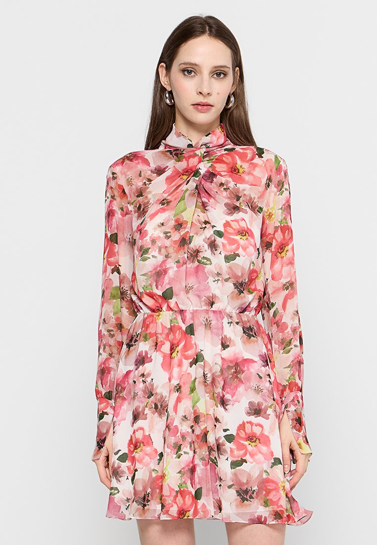 Vrouw in een lange mouw bloemenjurk met roze, rode en groene bloemen, staande tegen een effen lichte achtergrond.