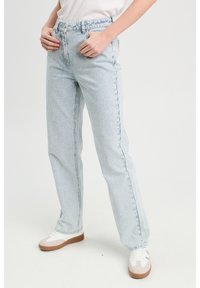 Jeans rayés bleu clair avec une taille haute, une coupe décontractée et des coutures visibles. Associés à des baskets blanches avec des accents bruns.