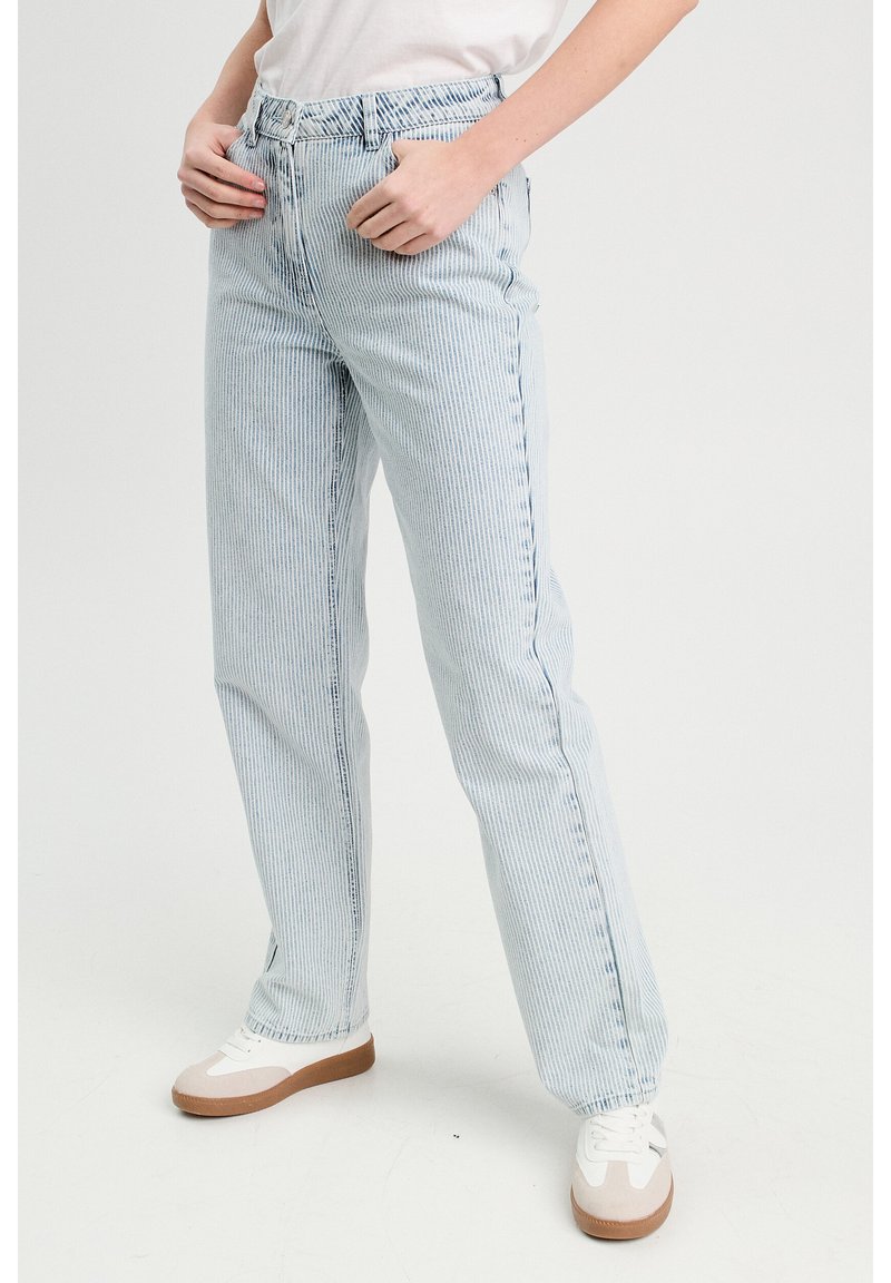Jeans rayés bleu clair avec une taille haute, une coupe décontractée et des coutures visibles. Associés à des baskets blanches avec des accents bruns.