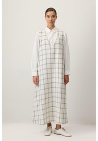 Robe à carreaux blancs avec motif à grille bleue, col en V, sans manches, portée par-dessus une chemise blanche à boutons, associée à des baskets blanches.