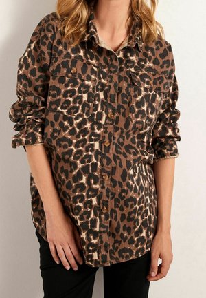 Femme portant une chemise à manches longues à boutons avec imprimé léopard marron et noir, et un pantalon noir, debout devant un fond clair.