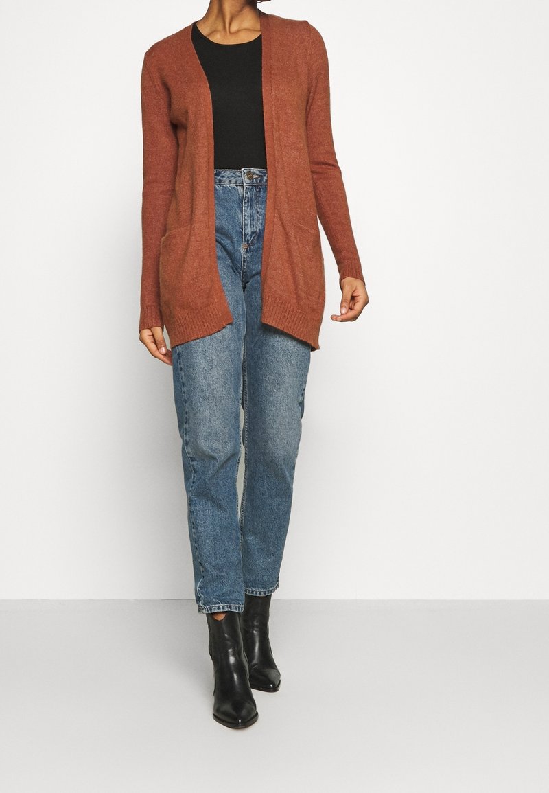 Cardigan couleur rouille en matériau doux, avec un devant ouvert, des manches longues et des poches. Associé à un jean taille haute bleu clair et des bottes noires.