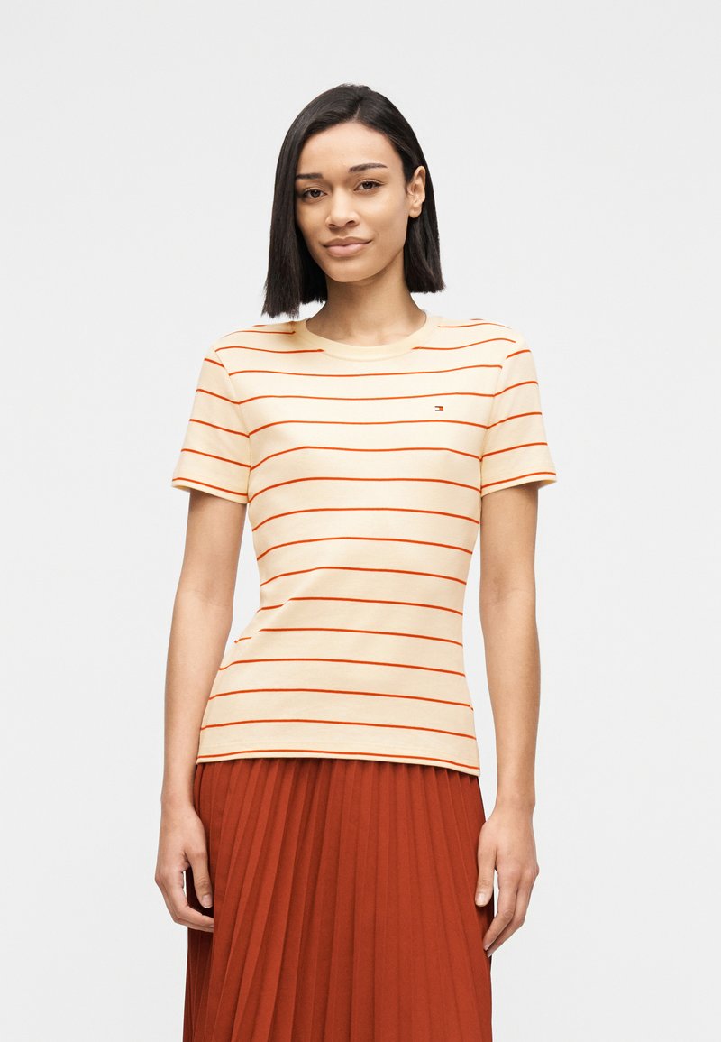 Γυναίκα με ίσια μαύρα μαλλιά, που φοράει κρεμ μπλούζα με λεπτές κόκκινες ρίγες και σκουρόχρωμη πιετέ φούστα, στέκεται μπροστά σε ένα απλό ανοιχτόχρωμο φόντο.