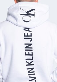 Sudadera con capucha blanca con un gran logo negro "CK" en la capucha y "CALVIN KLEIN JEANS" impreso verticalmente en la espalda con letras negras en negrita.
