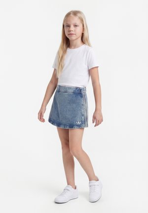 Ung pige stående iført en hvid t-shirt, blå denim nederdel med logo og hvide sneakers på en hvid baggrund.