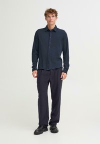 Marineblå knapp-up skjorte med lange ermer, flat krage og en subtil tekstur, parret med mørke bukser og svarte sko. En enkel, moderne design.