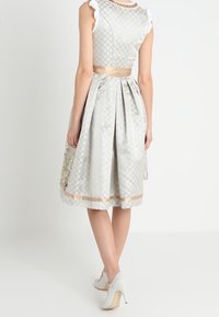Robe A-line argentée avec un motif texturé, manches blanches à volants, une ceinture en satin et des accents dorés à l'ourlet. Portée avec des talons blancs.