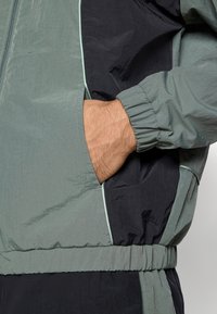 Kerge halli ja musta kangaga jakk, millel on elastsed mansetid, küljetaskud ja tõmblukk. Tekstuuriga materjal kontrastsete aktsentidega.