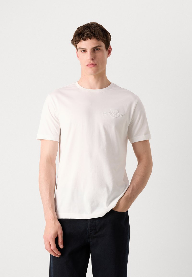 Baumwoll-T-Shirt mit Rundhalsausschnitt in hellbeige und kurzen Ärmeln, ausgestattet mit einem dezenten geprägten Logo auf der Brust, kombiniert mit dunklen Hosen.