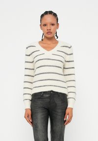 Pull en V en tricot de couleur crème avec des rayures grises. Il a des manches longues et un ourlet côtelé. Associé à un jean ajusté noir en simili cuir.