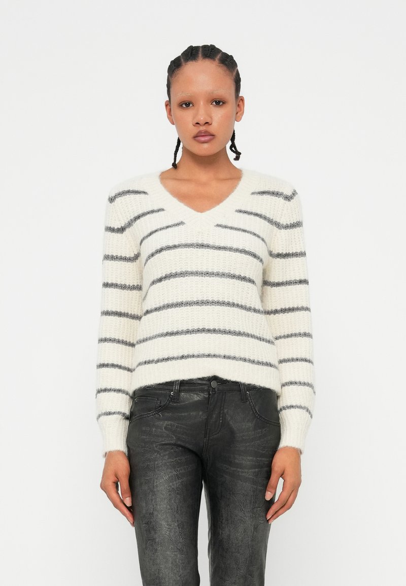 Pull en V en tricot de couleur crème avec des rayures grises. Il a des manches longues et un ourlet côtelé. Associé à un jean ajusté noir en simili cuir.