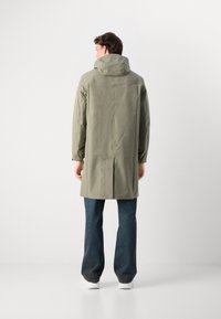 Parka impermeable verde claro con capucha, con mangas largas, cierre de botón en la parte trasera y puños elásticos, combinada con pantalones vaqueros oscuros.