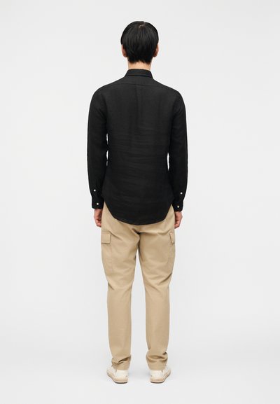 Bărbat stând cu spatele la cameră, purtând o cămașă neagră cu mâneci lungi, pantaloni cargo bej și pantofi albi, pe un fundal alb simplu.