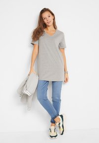 T-shirt grigia a V realizzata in tessuto morbido, abbinata a jeans blu e sneakers multicolori. Una giacca grigia chiaro è appoggiata su un braccio.