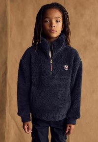 Pull-over en polaire marine avec col montant, ouverture zippée mi-hauteur et poignets côtelés. Présente un petit patch logo sur la poitrine. Tissu doux et texturé.
