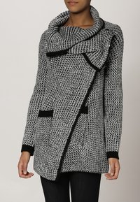 Cárdigan de punto asimétrico en blanco y negro con cuello amplio, mangas largas y bolsillos frontales; cuenta con detalles de ribete negro.