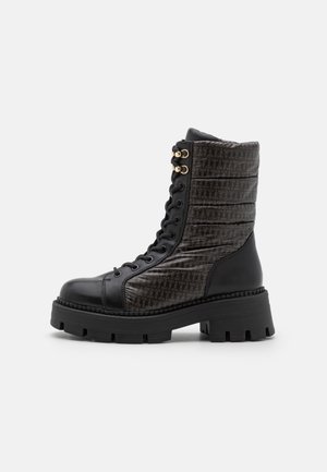 Bottine noire à lacets avec une tige synthétique texturée, présentant un panneau à motif sombre et une semelle en caoutchouc épaisse. Accents en métal doré.