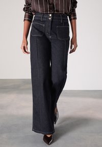 Jeans en denim foncé à jambes larges avec taille haute, deux poches avant, coutures contrastées et fermeture par deux boutons métalliques.