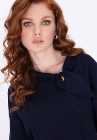 Top tricotat de culoare bleumarin, cu o accentuare mare sub formă de fundă la gât, având un detaliu cu nasture aurie. Textură moale, design ajustat și mâneci până la trei sferturi.