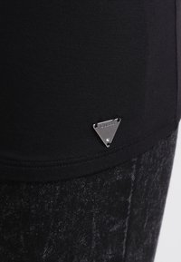 Musta kangas, jossa on pieni hopeinen kolmion muotoinen metallilogo, johon on kaiverrettu "GUESS", kiinnitetty vaatekappaleen helmaan.