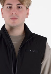 Gilet nero con zip e colletto alto, con un piccolo logo Calvin Klein. Indossato sopra un maglione beige chiaro con un logo simile.